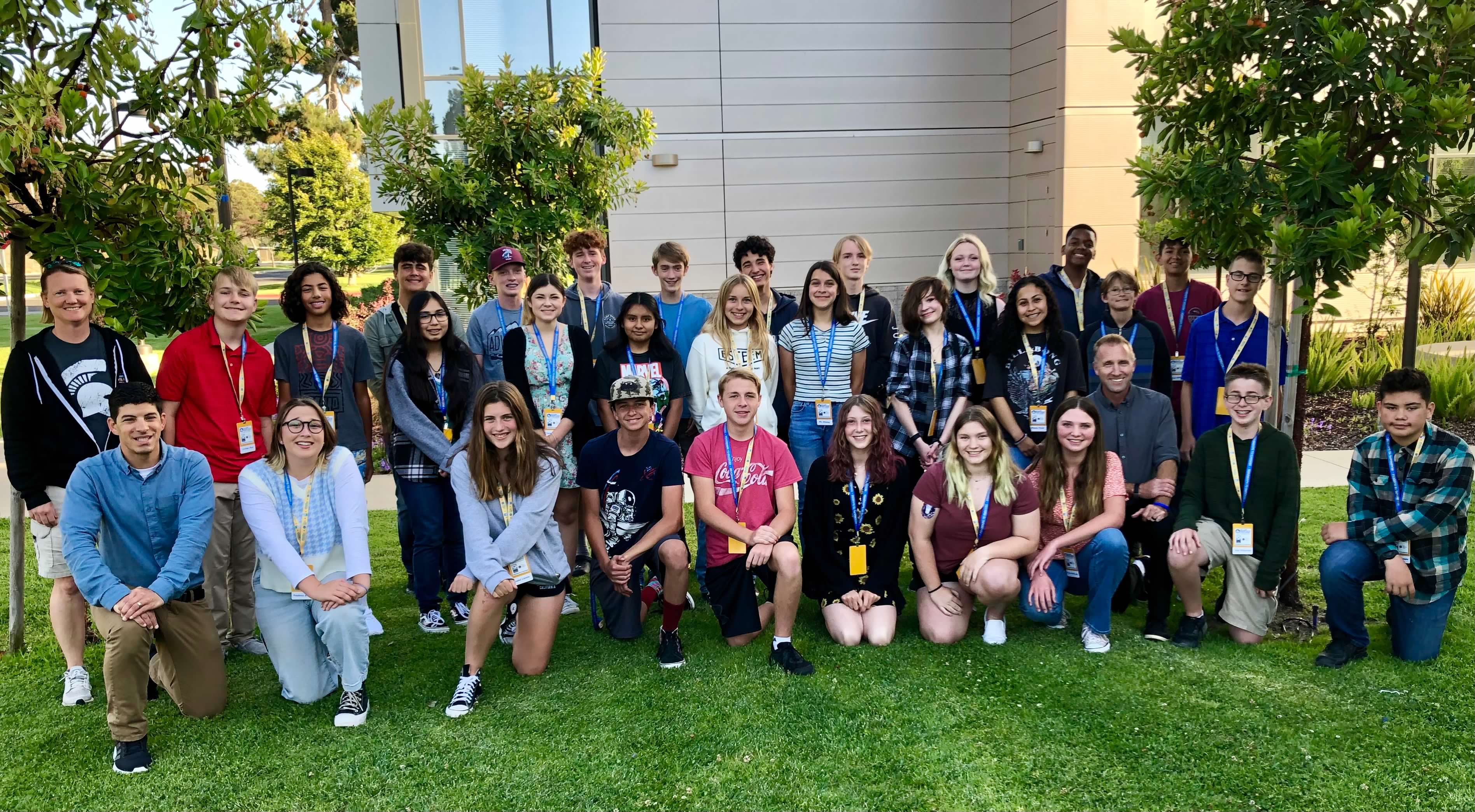 Group photo of students participating in Orcutt Academy High School’s Early College Cadre program. The students and staff stand and kneel together on a grassy campus area outside a college building, wearing name badges and smiling for the camera during the academic program event.